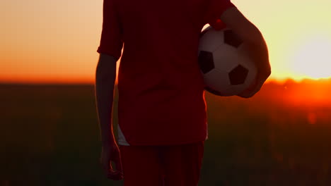 Ein-Kleiner-Fußballspieler-In-Einem-Roten-T-Shirt-Steht-Mit-Einem-Fußball-Bei-Sonnenuntergang-Auf-Dem-Feld